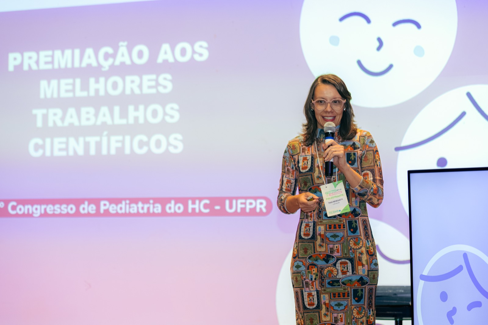 Palestrante do 1º Congresso de Pediatria do HC-UFPR no palco, ao lado do telão da premiação aos melhores trabalhos científicos